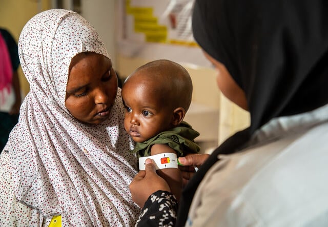 A mother holds her young child, who has their arm measured by an IRC nutrition officer as part of a screening for signs of malnutrition.