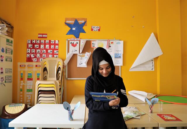 A young girl smiles down at a book that she is reading in a classroom, which features bright yellow walls in the background.