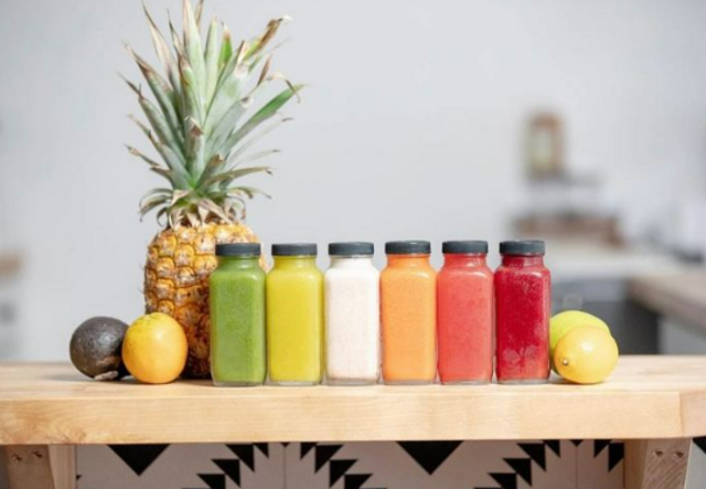 A row of multi-colored bottles of Yogi Elixir juices sitting on a table alongside an avocado, two lemons, a lime, and a pineapple.