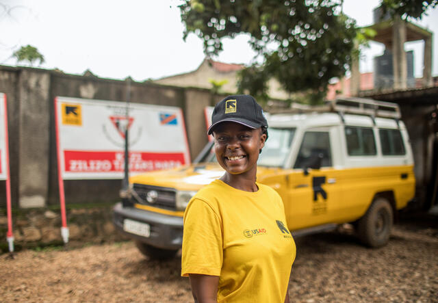 An IRC staff member smiles and poses for a pictures outdoors, and in front of a car that features SUV branding.