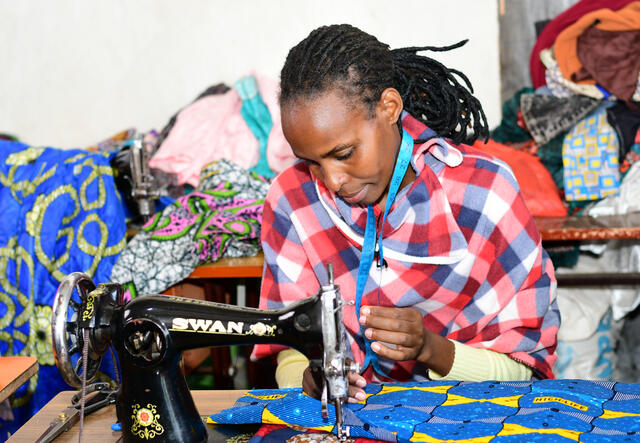 A woman sits at a desk with a sewing machine and works intently on a piece of clothing. Behind her lay other pieces of clothing.