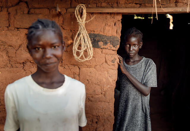 Two sisters pose for a photo outside of a clay building in South Sudan. One sisters stands in the foreground while the other a few feet behind her.