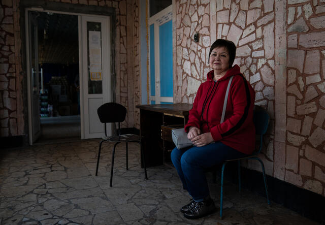 Larysa poses for a photo outside a building in Ukraine. She sits on a chair and sports a faint smile and poses for a photo after receiving cash assistance from the IRC.