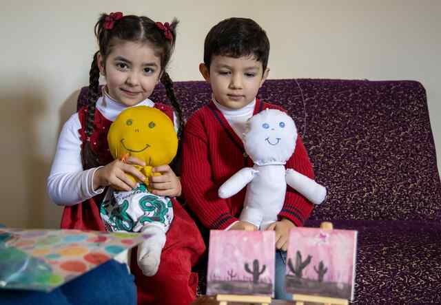 Two young children, Mira and Jawad, sit on a couch and proudly hold dolls that they made during an Ahlan Simsim program.
