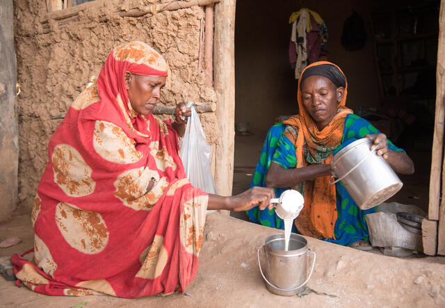 Two women sit on the ground in a Kenyan village and begin to make food using maize powder and milk.