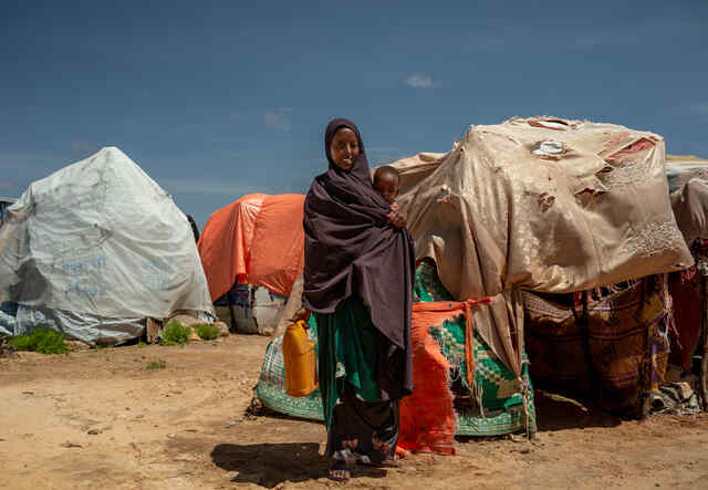Halima stands in front of her home with her baby in her arm.