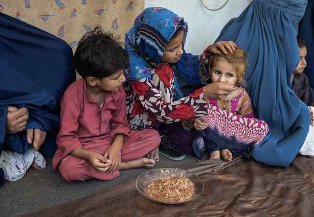 A mother sits with two children and feeds one of them from a bowl of food.