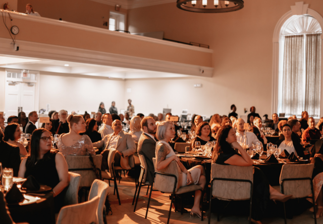 An audience of Gala attendees sitting at their dinner tables and their attention affixed to a presentation out of frame.