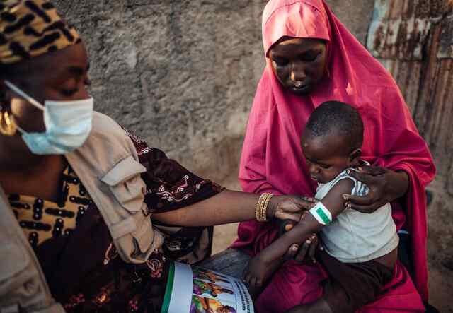 An IRC nutrition officer teaches a mother how to screen her child for signs of malnutrition using MUAC tape in Nigeria. The malnutrition wraps the tape around a boys arm while the mother looks on.