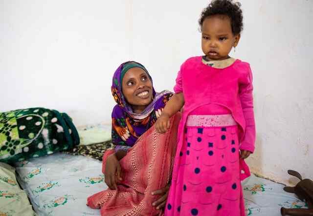 Iftu looks up with a big smile at her daughter, Fidiya, who stands wearing a pink dress.