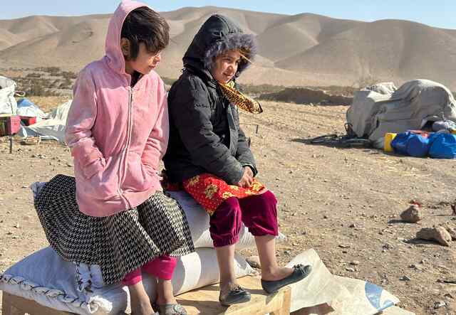 Zwei Mädchen sitzen auf einer kleinen Bank im Freien. Im Hintergrund ist die Landschaft Afghanistans mit Hilfsgütern für die Erdbebenhilfe übersät.