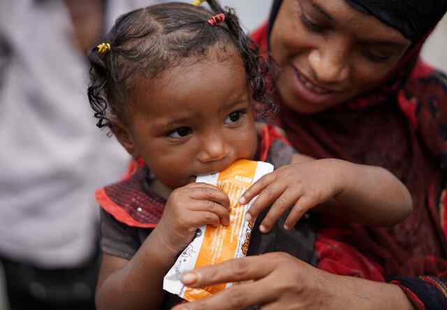 A young child is held by a caregiver while they eat a healthy peanut paste, provided by the IRC.
