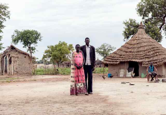 A man and woman poses for a photo outside their home in South Sudan. Behind the man, a boy sits on a chair.