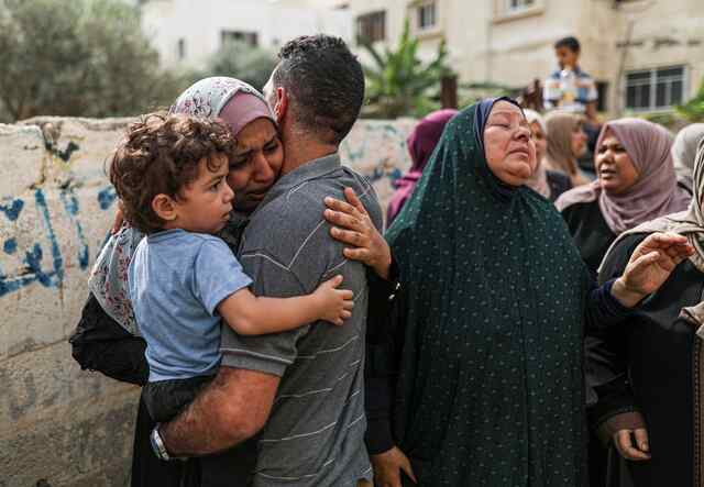 A family embraces each other with looks of distress on their faces. In the foreground, a man holds a small child who looks on.