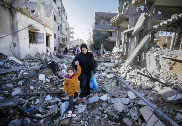 A young child wearing a yellow jacket holds an adult's hands while they walk through a destroyed neighborhood in Gaza.