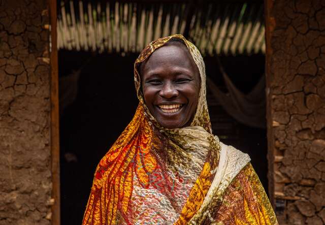 A woman sports a wide smile as she poses for a portrait outside of a building in South Sudan. She is wearing a colorful headscarf.