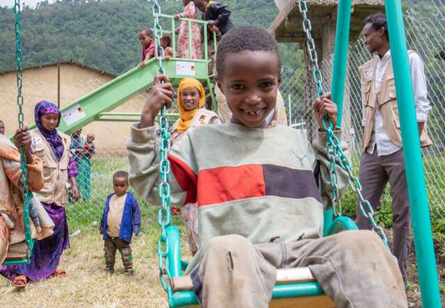 An 8 year old boy smiles as he swings on a swing-set at an IRC child protection center in Ethiopia. Children play alongside IRC staff in the background.