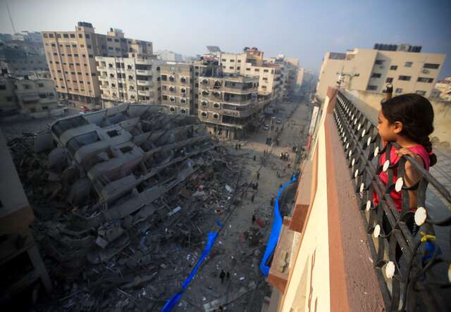 A young Palestinian girl stands on a rooftop, looking down upon the ruins of a destroyed building.