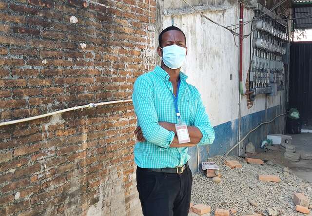 A man in a medical masks and a blue shirt stands against a building, posing for a picture.