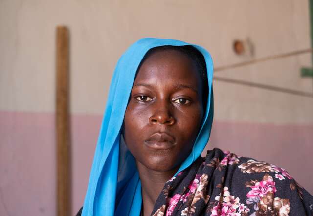 Portrait of a woman staring solemnly into the camera. She wears a blue headscarf.