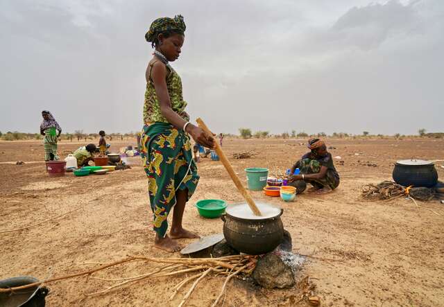 A girl stirs a cauldron outside in Burkina Faso. Behind her, other women cook food.