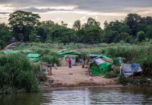 Myanma refugees stand in a makeshift refugee camp near a river.