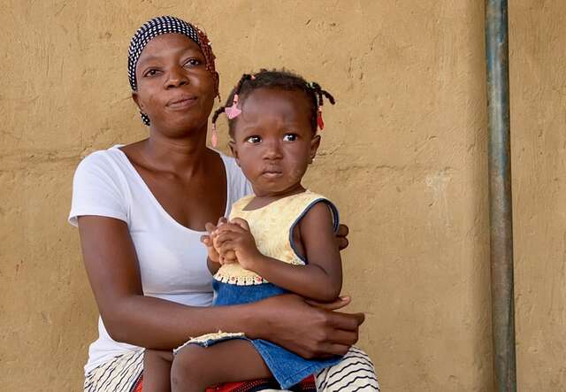 A mother holds her daughter in her lap. Both of them look towards the camera.