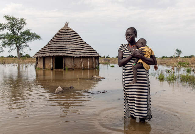 En mamma i Sydsudan håller sitt barn framför deras översvämmade hus.