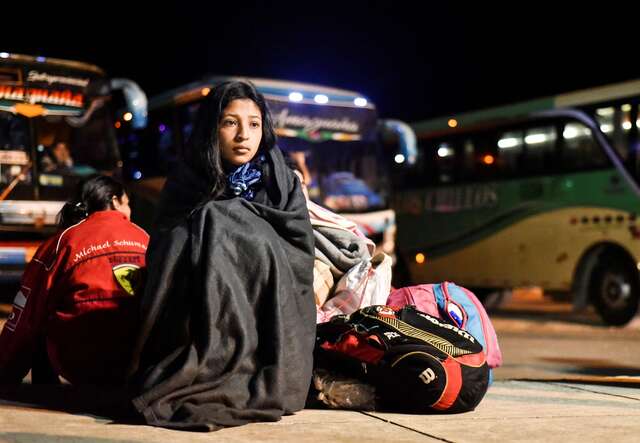 A young woman sits at a bus depot, wrapped in a blanket, next to several bags.