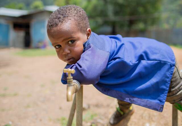 A young boy wearing a blue shirt leans over a water pipe and poses for a photo.