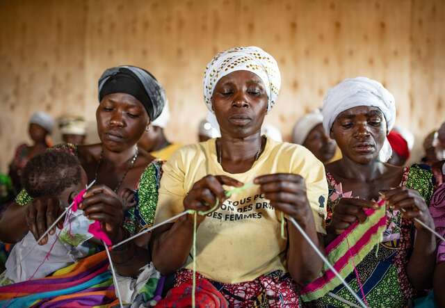 Three woman sit together and knit. One of them holds a young child in her arms.