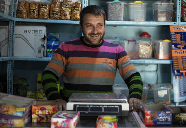 Mann in einem bunt gestreiften Pullover steht hinter einer Theke in einem Geschäft, umgeben von verschiedenen Produkten.