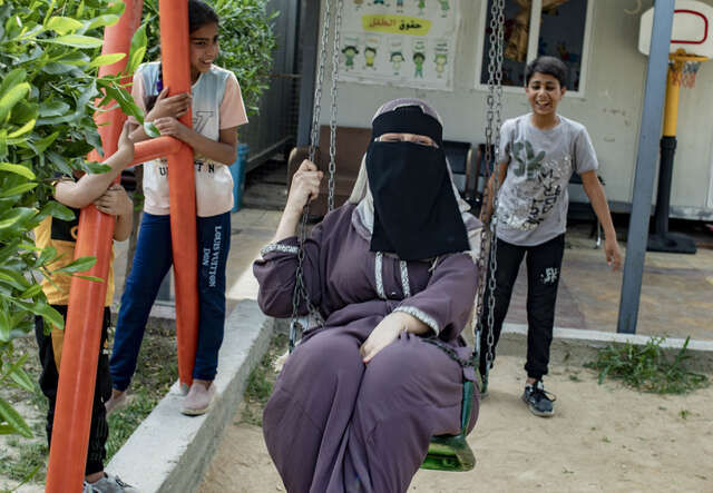 Eine Person schaukelt in einem Außenbereich, während zwei weitere Personen daneben stehen und die Szene beobachten.
