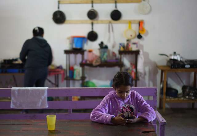 A nine year old girl sits at a table in a shelter in Juárez, Mexico. Her mother cooks food in the background.
