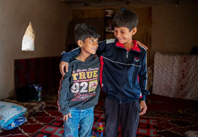 Two brothers smile at each other, with one arm wrapped around each other; They are standing in their grandparents humble home in an informal settlement for internally displaced Syrians.