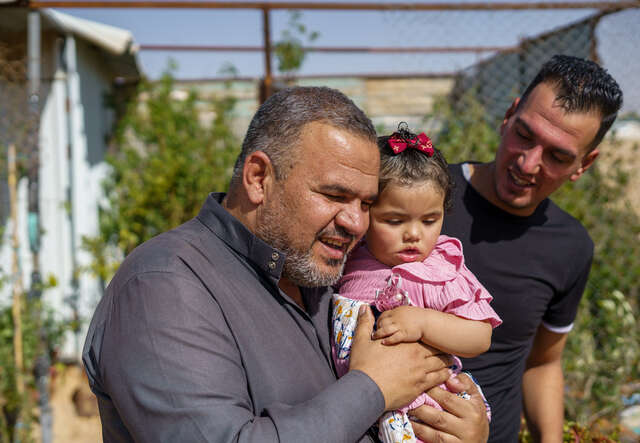 En man i Jordanien kramar sitt 1-åriga barnbarn. Bredvid dem står flickans pappa. Familjen har flytt från Syrien.
