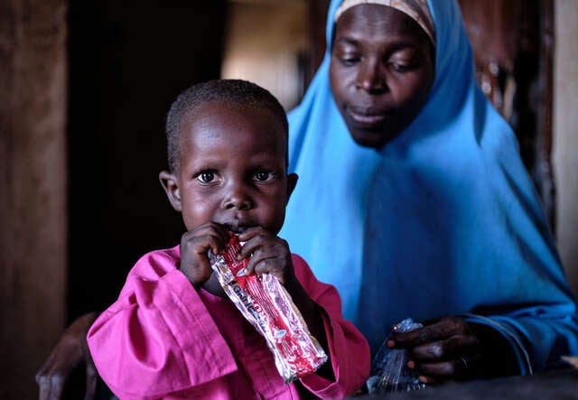 En liten pojke sitter i sin mammas knä och håller i en nötkrämspåse, han får behandling mot undernäring i ett flyktingläger i Nigeria.
