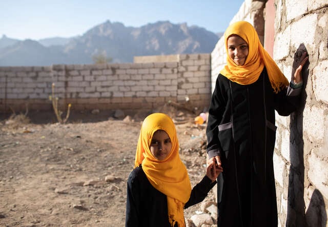 Two young girls, wearing matching headscarves, hold hands by a wall in Yemen.