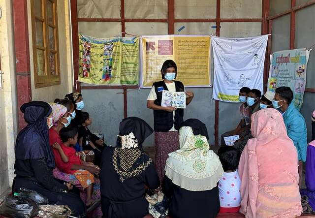 A person is holding a card in front of a group of adults and children wearing face masks.