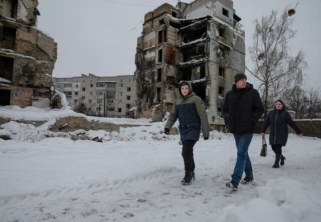 Three Ukrainians walk past the ruins of an apartment building that has been destroyed by a missile strike. The area is cold and covered in snow.