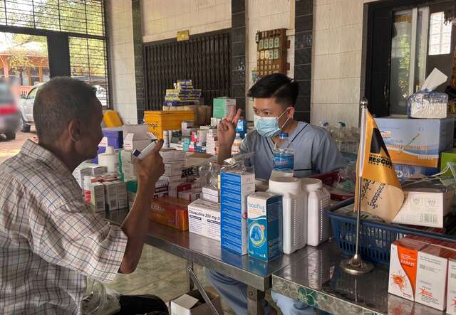 A man with a mask gestures to a pharmacist behind the counter with medicine supplies from the IRC