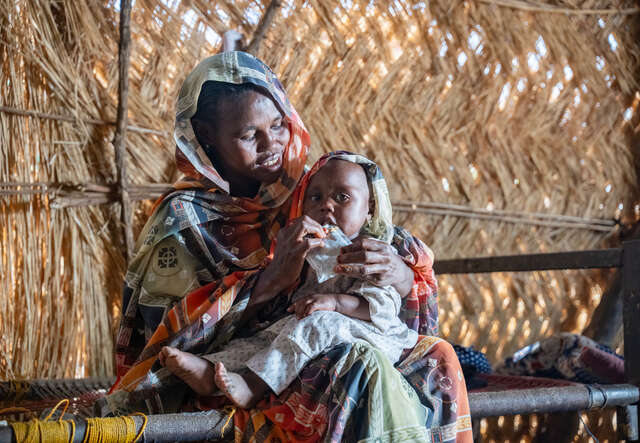 En pojke sitter i sin mammas knä och matas med nötkräm.