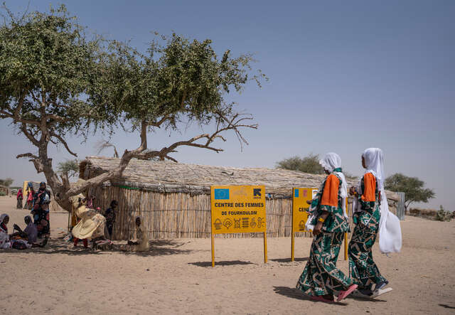 Två flickor går mot en samling av kvinnor som sitter i skuggan av ett träd.