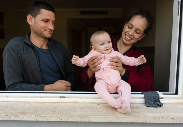 En pappa och mamma håller i sin fem månader gamla dotter vid ett fönster.