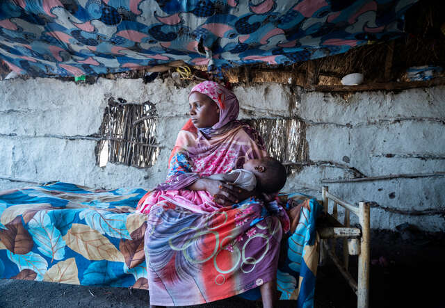 En mamma sitter på en säng i ett skjul och håller sin son i famnen. 