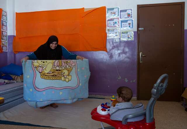 Warda folds a blanket while her baby, Mohammad, looks on.