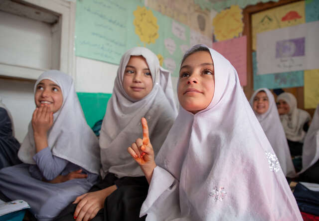 Tre flickor i Afghanistan sitter i ett klassrum och tittar mot sin lärare, en av dem räcker upp handen.