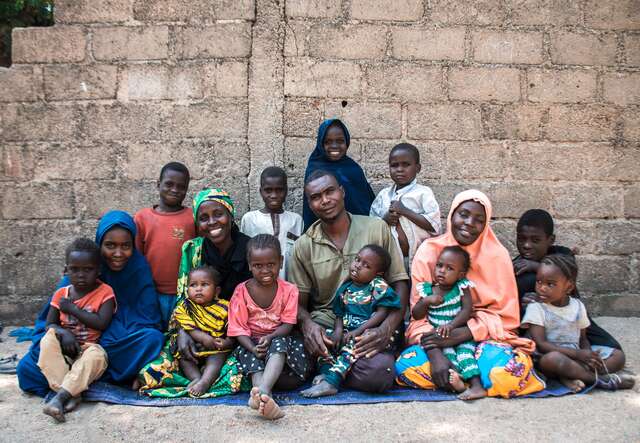 Abubakar and his wives, Hawa (in green on the left) and Zara (in pink on the right), and eleven of their children seated on the ground to take a family portrait.