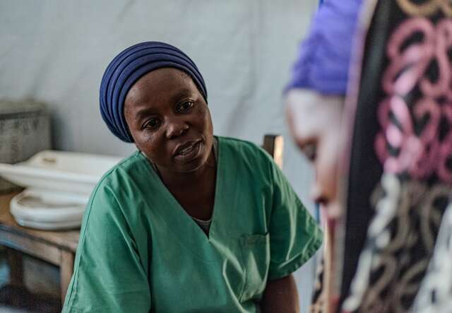 Neema, a midwife wearing green scrubs and a blue headwrap, speaks with a patient at the Numbi health center in South Kivu, Democratic Republic of Congo.