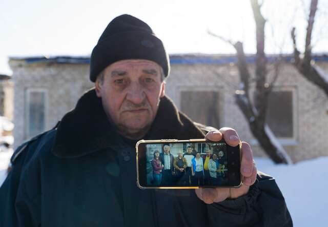 An elderly man holds up a picture of his family in front of a house, on his mobile phone. The background is snowy.